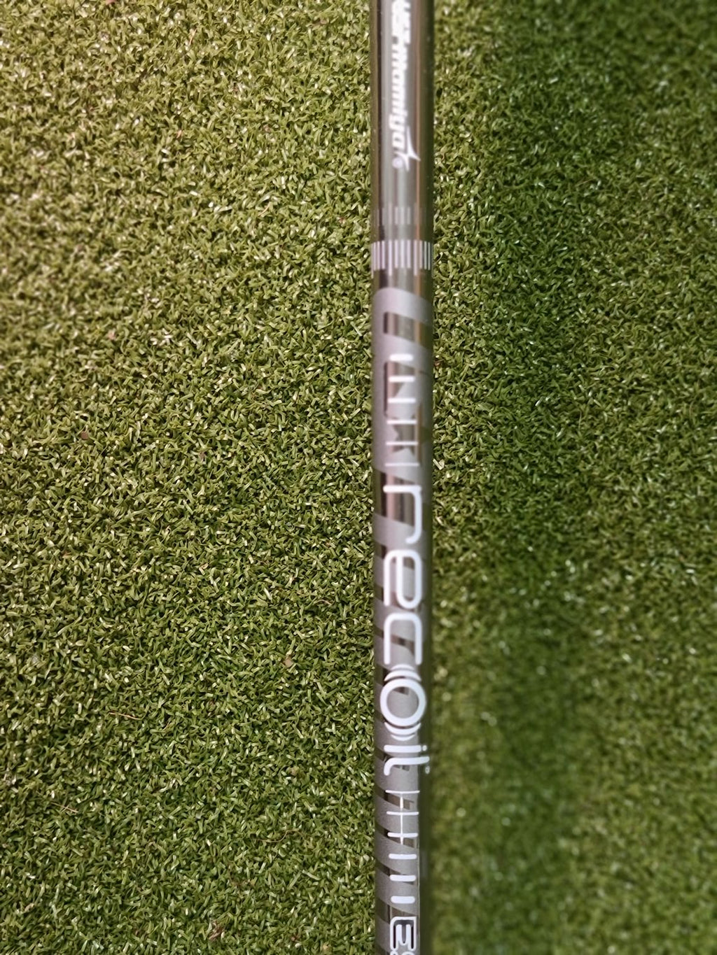 A close-up of a golf shaft resting horizontally on green artificial turf. The shaft features the 'Mizuno' brand name and 'NECO' model text in white and silver lettering.