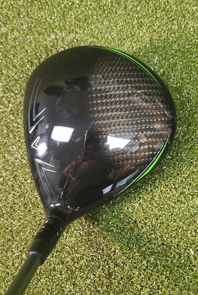 A carbon fiber golf driver head with a black finish and a thin green accent line, resting on green artificial turf.