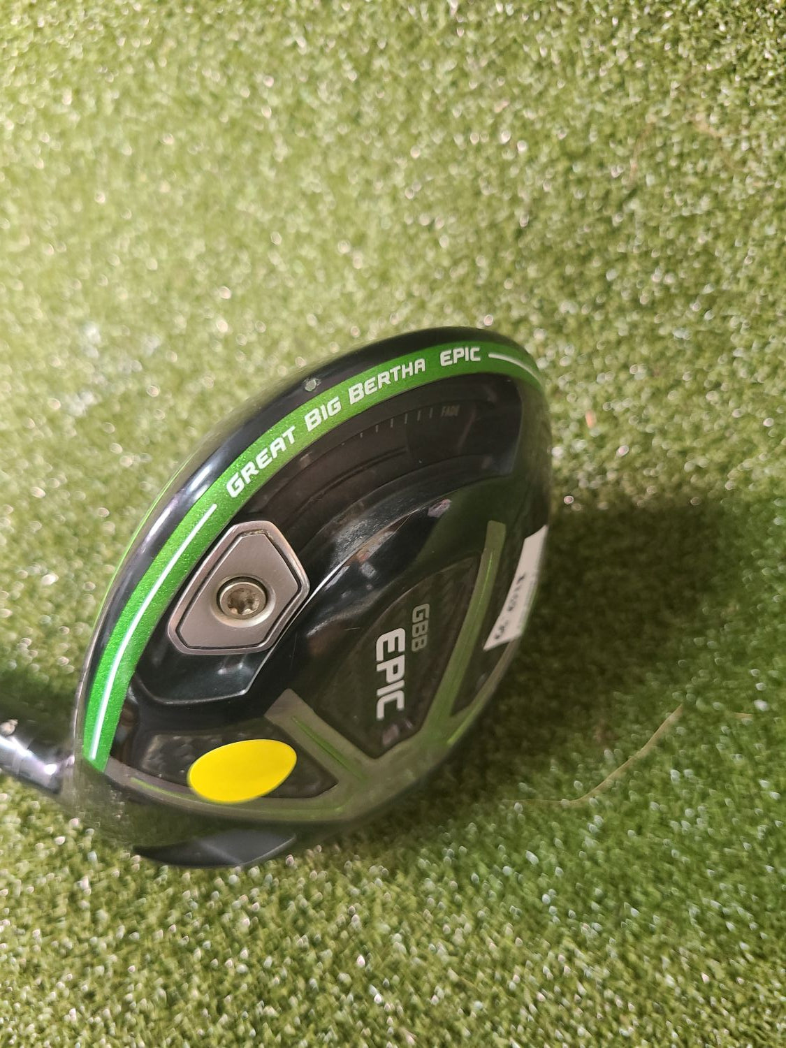 A Callaway Epic golf driver club head on a green turf background, featuring a black and dark gray finish with green accents, white lettering, and a yellow button.