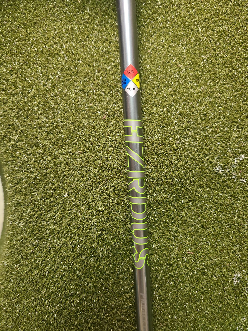 A close-up of a golf shaft labeled 'HIZRIDUS' in neon yellow-green lettering, resting on artificial green turf. The shaft is dark gray with a colorful logo sticker near the left end.