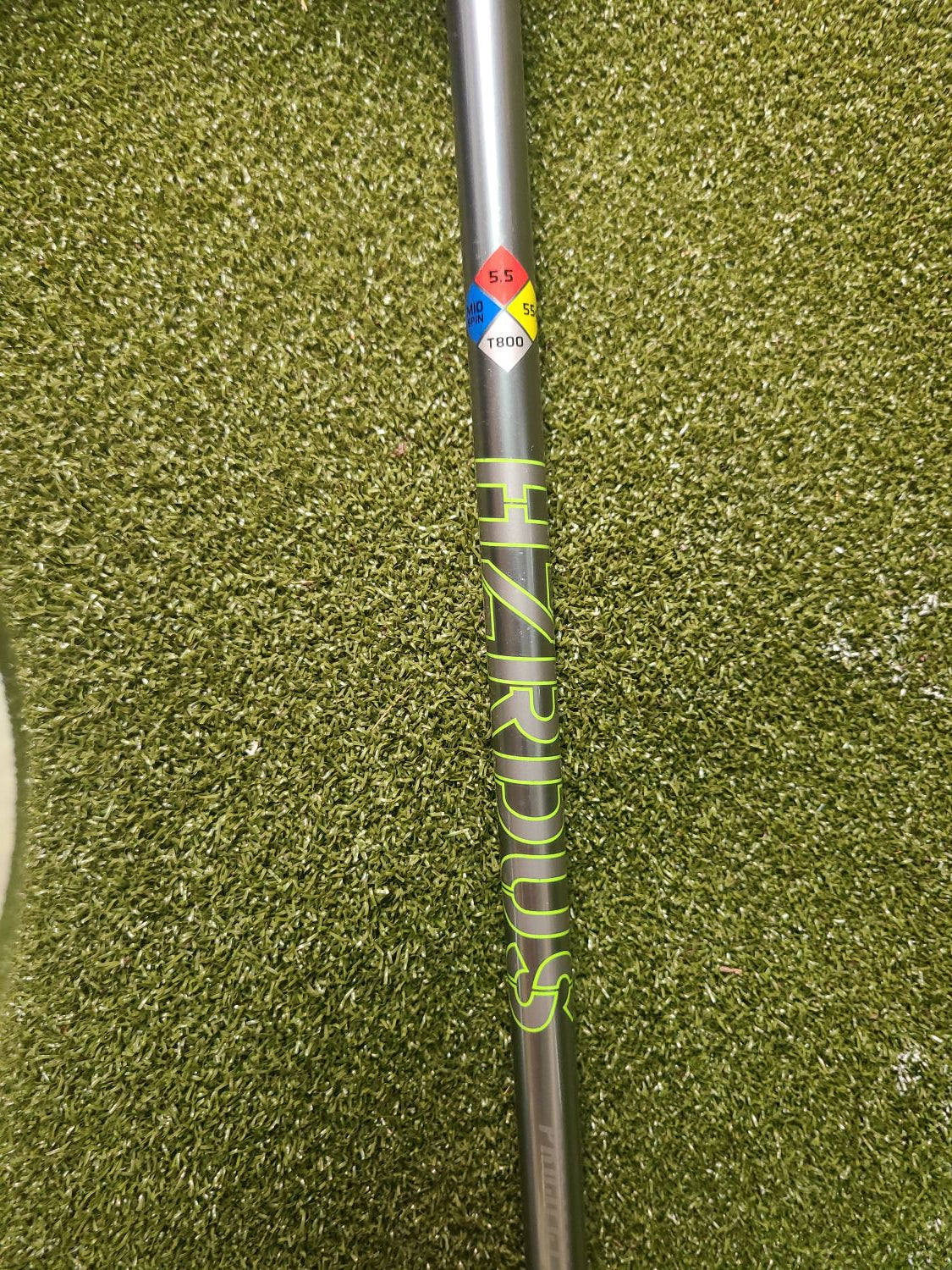 A close-up of a golf shaft labeled 'HIZRIDUS' in neon yellow-green lettering, resting on artificial green turf. The shaft is dark gray with a colorful logo sticker near the left end.