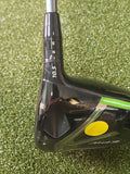 Close-up of a black and green golf driver club head with a yellow sticker, resting on green artificial turf.